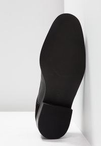 Chaussure en suède noir avec un dessus lisse et texturé et une semelle plate en caoutchouc, présentant des rainures horizontales pour une meilleure adhérence.