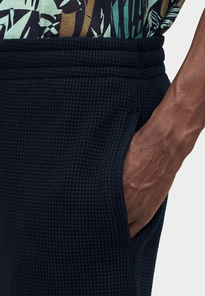 Χέρι στην τσέπη σκοτεινού υφασμάτινου παντελονιού με ελαστική μέση, φοράει πουκάμισο με πράσινα, μαύρα και καφέ σχέδια.