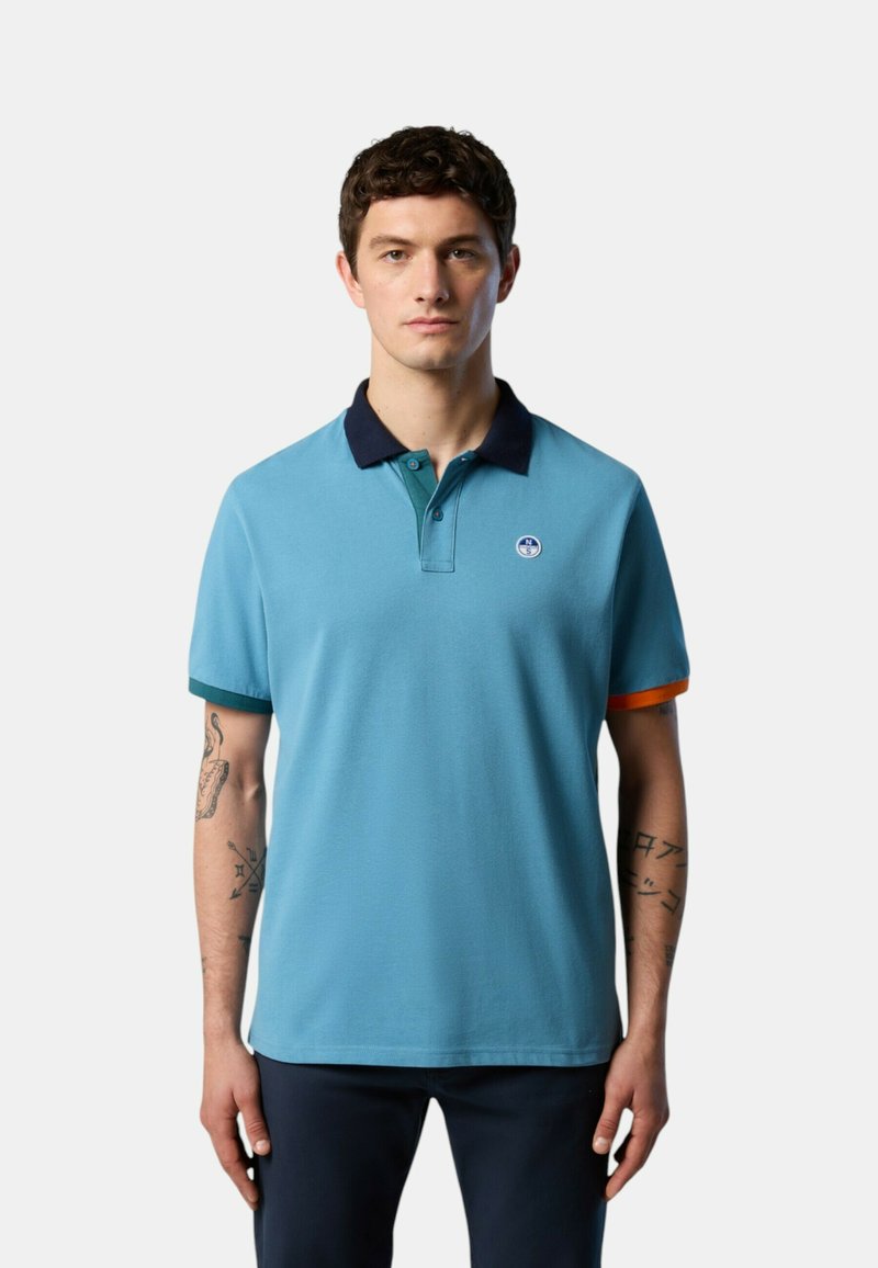 Polo shirt azzurro chiaro con colletto blu scuro, dettagli arancioni sulle maniche e un logo circolare sul petto. Tessuto di cotone liscio.
