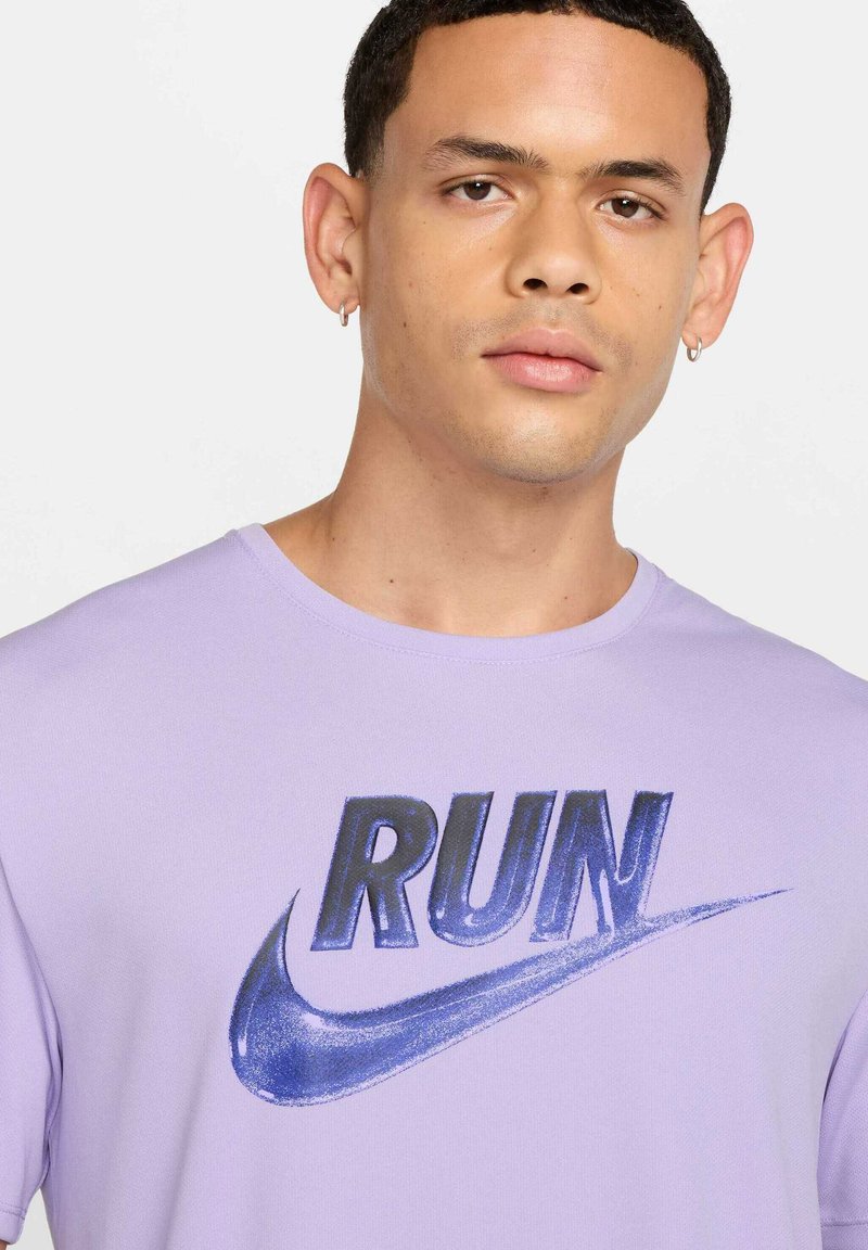 Jeune homme aux cheveux courts portant des boucles d'oreilles argentées en forme d'anneaux et une chemise violet clair avec l'inscription "RUN" et un logo Nike en forme de virgule bleu.