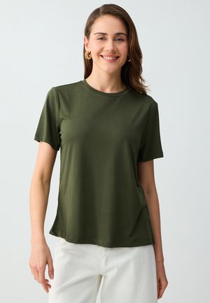 Donna sorridente con capelli castani di media lunghezza indossa orecchini a cerchio dorati, una t-shirt verde oliva a maniche corte e pantaloni bianchi, in piedi davanti a uno sfondo semplice.