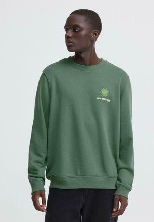 Mann trägt ein grünes Sweatshirt mit einer Sonnen-Grafik und dem Schriftzug "SUN SEEKERS", schaut nach links vor einem einfarbigen hellen Hintergrund.