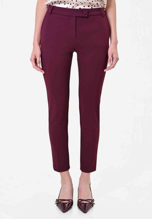 Femme debout portant un pantalon ajusté violet foncé longueur cheville, une blouse à pois et des talons brillants bordeaux à bout pointu avec des nœuds.