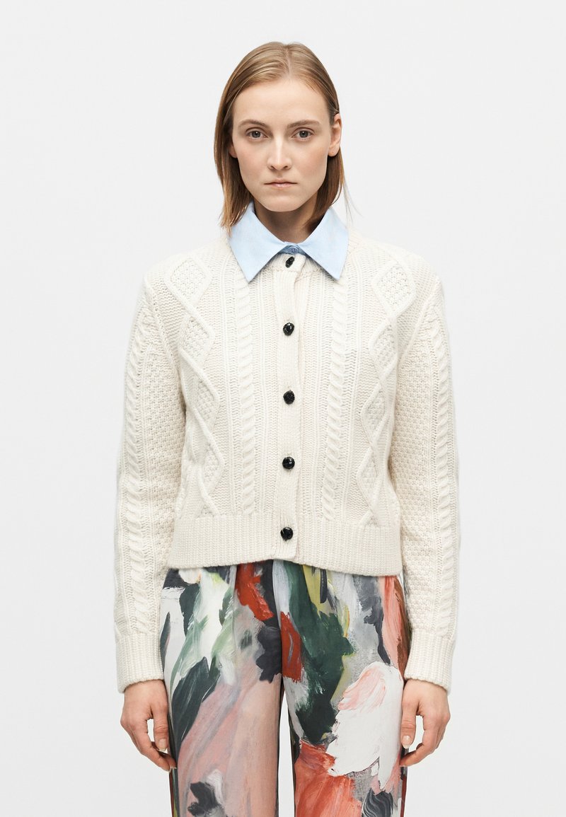 Cardigan crème en maille torsadée avec boutons noirs, coupe courte et ourlet côtelé. Superposé avec une chemise à col bleu clair en dessous.