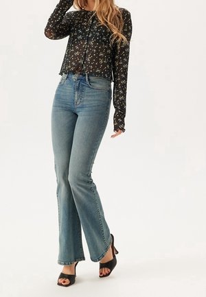 Mujer vestida con vaqueros azules de talle alto y pata de elefante, blusa negra de manga larga con estampado floral y sandalias negras de tacón con punta abierta, de pie sobre fondo blanco.