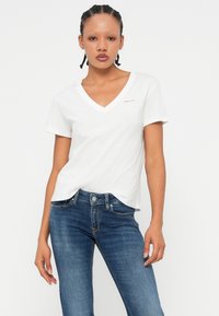 T-shirt bianca a maniche corte con scollatura a V, realizzata in cotone leggero, con un piccolo logo rosso sulla parte sinistra del petto, abbinata a jeans blu.