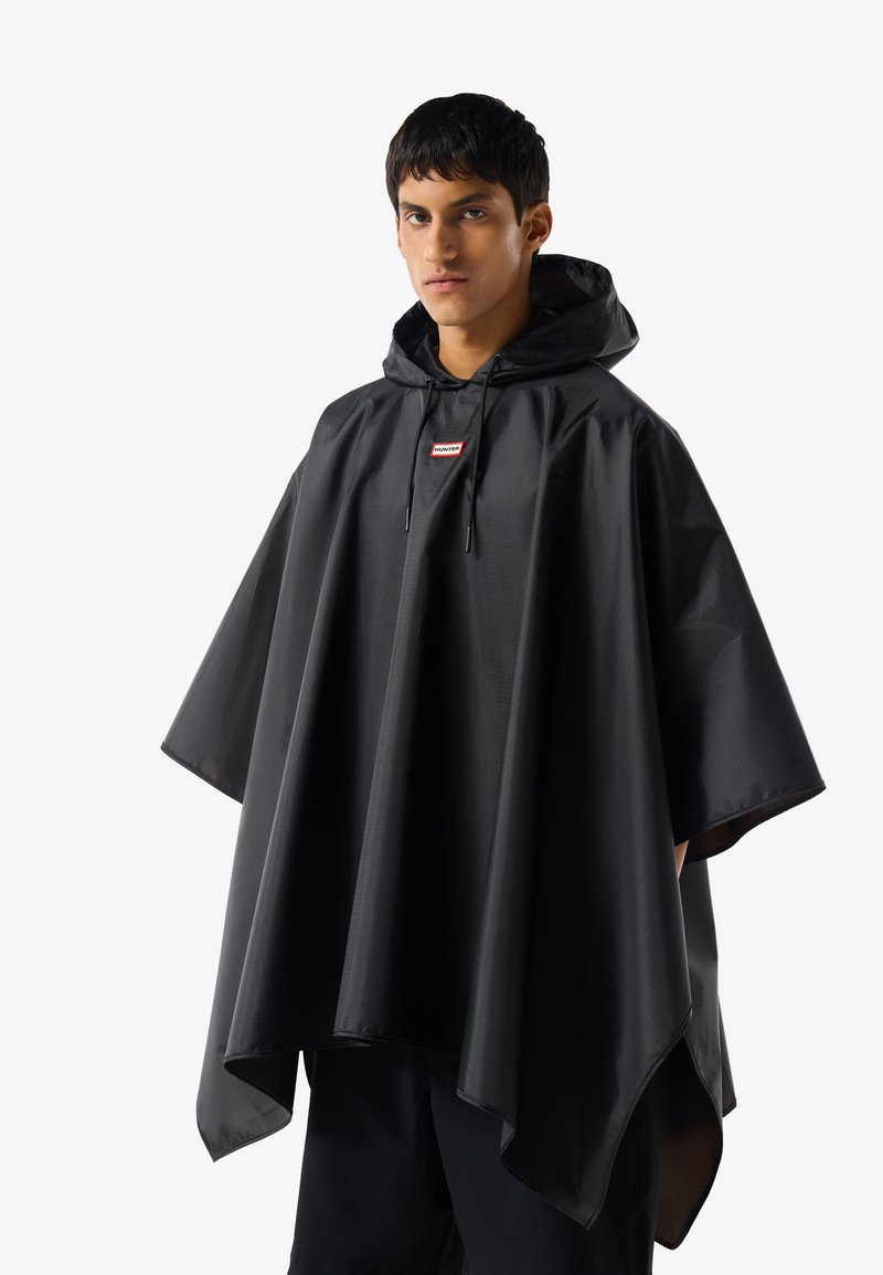 Νεαρός άνδρας που φοράει μαύρο αδιάβροχο πανωφόρι με κουκούλα και μικρό κόκκινο λογότυπο στο στήθος, στέκεται μπροστά σε απλό φόντο.