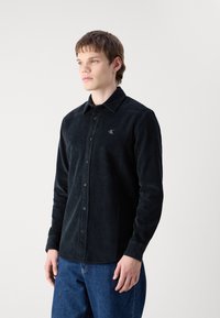 Camicia button-up in corduroy nera con colletto e logo ricamato sottile, abbinata a jeans di denim blu, indossata da un modello.