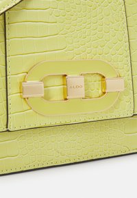 Limegrön handväska med krokodilmönster, med ett guldtonat lås med "ALDO"-logotyp och kontrasterande svart kant.