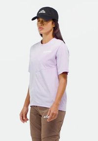 Svetlofialová košeľa s krátkym rukávom a okrúhlym výstrihom, s detailom na prednom vrecku; skombinovaná s hnedými nohavicami s logom. Čierna šiltovka dokončuje outfit.