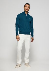Teal zip-neck tröja med ribbad ärm- och nederkant, tillsammans med vita byxor och ljusbeige sneakers. Enkel logotypdetalj på tröjan.