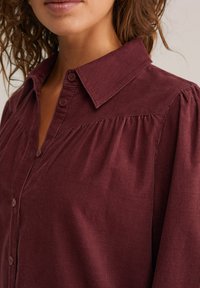 Burgundi värvi korduroy särk, millel on nööpidega esikülg, suur krae ja pintuck-üksikasjad õlgadel, näidates pehmet tekstuuriga kangast.