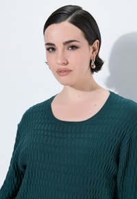 Ung kvinde med kort mørkt hår, iført guldøreringe og en struktureret mørkegrøn sweater, poserer foran en helt hvid baggrund.