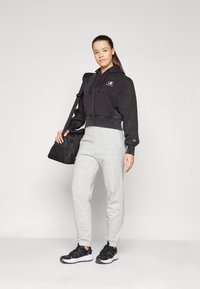 Svart avskåret hettejakke med logo, grå joggebukser, svart og hvite joggesko. Modell som bærer en svart duffelbag. Enkel, atletisk stil.