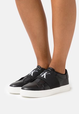 Jambes portant des baskets plates en cuir noir avec de larges bandes élastiques affichant le logo "cK" blanc et des semelles blanches épaisses.