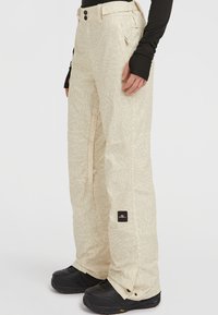Beige sneeuwbroek met een subtiel abstract patroon, vastgehouden met twee knoppen en met zakken. Draagt zwarte sneeuwlaarzen.