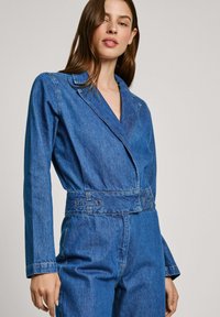 Denim kombinezon z dolgimi rokavi, zarezalnim ovratnikom, zategnjenim pasom in stranskimi žepi. Srednje modra pranje z vidnimi šivi.