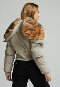Korte pufferjack in beige met een bruin nepbontkraag. Voorzien van een rits sluiting, geribbelde manchetten en een geborduurd logo op de achterkant.