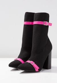 Mustat nilkkurit, joissa on neulottu päällinen, vaaleanpunaiset kiiltävät hihnat nilkassa ja varpaissa, joissa on hopeiset soljet, ja paksu korkea korko.