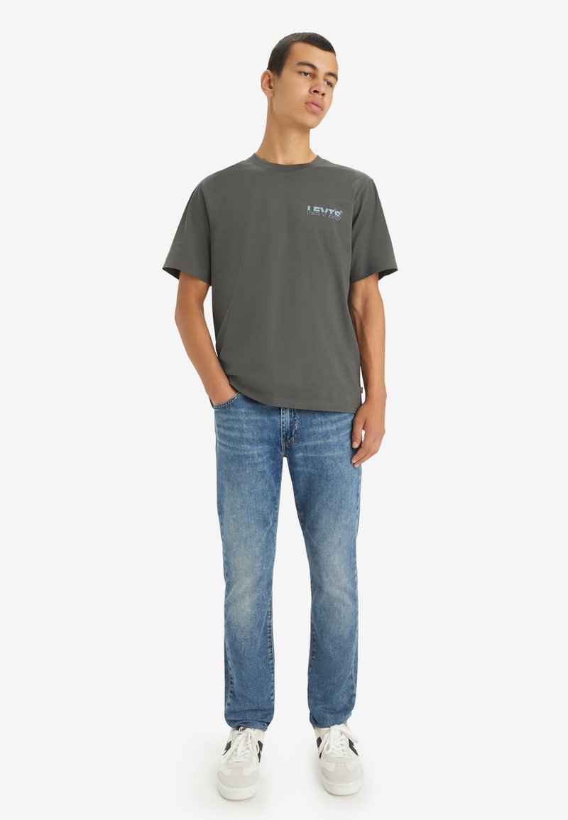 T-shirt gris à manches courtes avec le logo bleu "LEV's", associé à un jean bleu clair et des baskets blanches avec des accents noirs.