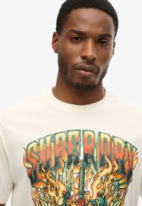 Camiseta de algodón color crema con un diseño gráfico colorido que incluye llamas, una espada y el texto "Superdry" en una fuente audaz y estilizada.