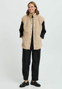 Beige, ärmellose Weste aus plüschigem, strukturiertem Stoff mit einem glatten Kragen; Druckknopfschluss und seitliche Taschen. Schwarze Hosen und Schuhe.