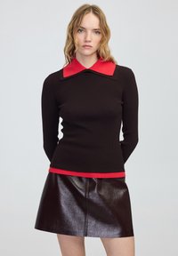 Młoda kobieta z falującymi blond włosami, ubrana w czarny sweter z prążkowaną fakturą, ozdobiony czerwonym kołnierzykiem i wykończeniem, w połączeniu z ciemną, błyszczącą skórzaną spódnicą.
