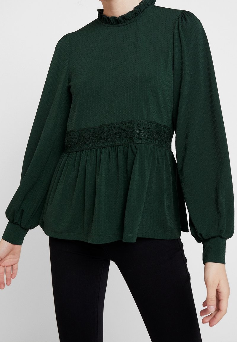 Zelená blůza s texturovanou tkaninou, vysokým volánkovým výstřihem, dlouhými rukávy a krajkovým pasem. Má rozšířený peplum styl.