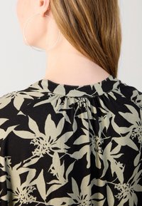 Gros plan sur le dos d'une femme aux cheveux longs châtain clair, portant un haut noir orné de grands motifs floraux beiges et une grande boucle d'oreille argentée en forme d'anneau.