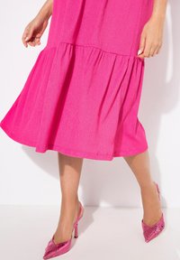 Vaaleanpunainen teksturoitu mekko, jossa on liehuva helma ja kerroksellinen röyhelö. Yhdistetty kiiltävän magentan värisiin slingback-korkokenkiin, joissa on teräväkärkinen muotoilu.