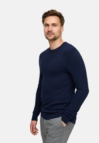 Granatowy sweter z dzianiny o teksturowanym wzorze w warkocze, okrągły dekolt oraz ściągacze przy mankietach i dolnym brzegu. Nosi się go z szarymi spodniami, charakteryzuje się slim fit.