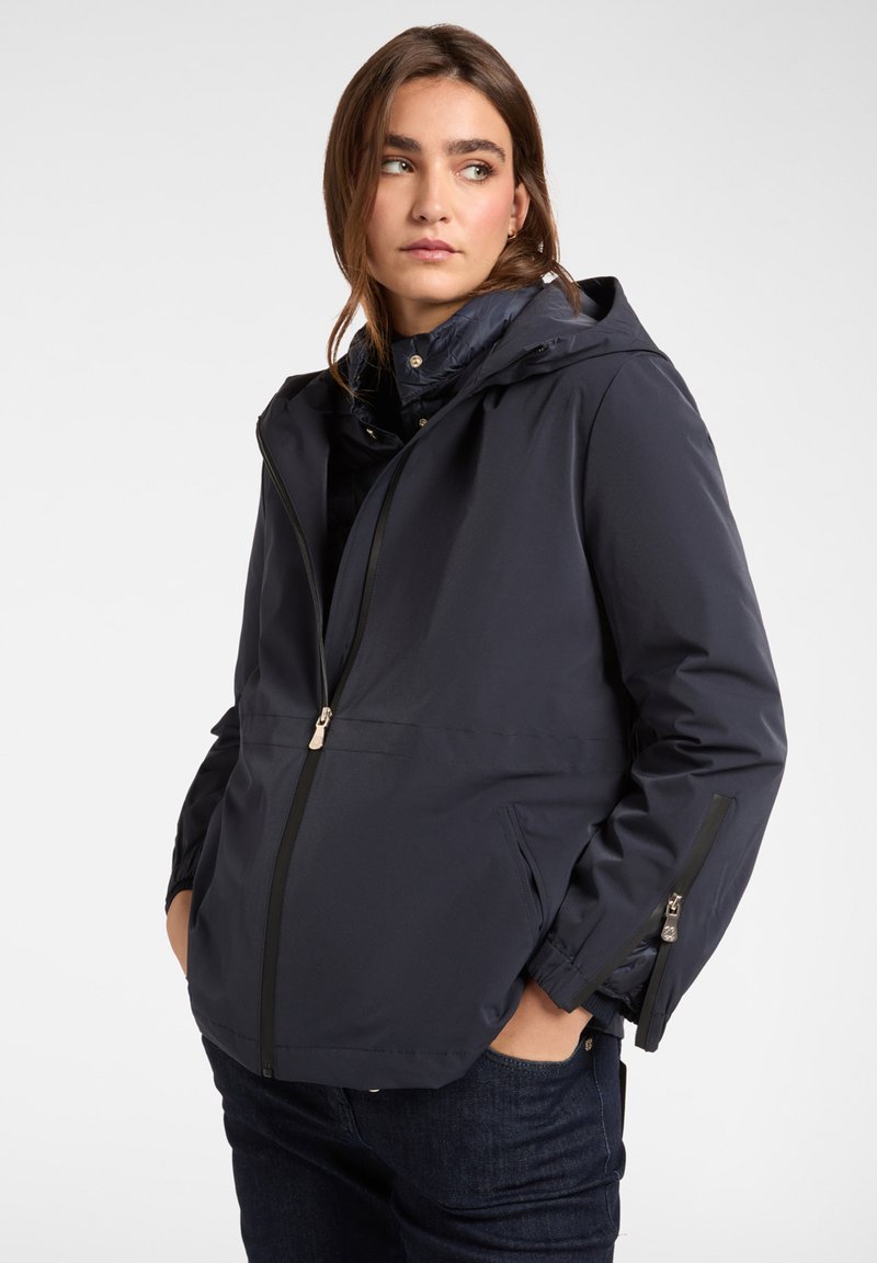 Giacca impermeabile blu navy con cappuccio, chiusura frontale con zip e tasche laterali; presenta una texture liscia e polsini regolabili.