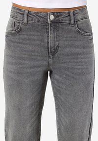 Blugi gri din denim cu talie înaltă, prevăzuți cu nasture frontal, fermoar, bucle pentru curea și buzunare frontale, purtați cu un top alb scurt.