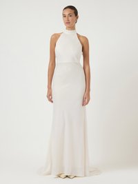 Γυναίκα ντυμένη με ένα μακρύ, χωρίς μανίκια λευκό φόρεμα με λαιμόκοψη halter, στέκεται με φόντο μια απλή, ανοιχτόχρωμη επιφάνεια.