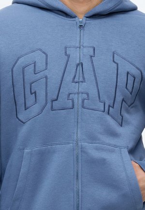Gros plan d'un sweat à capuche bleu zippé avec de grandes lettres en relief "GAP" sur la poitrine et une main partiellement dans la poche avant.