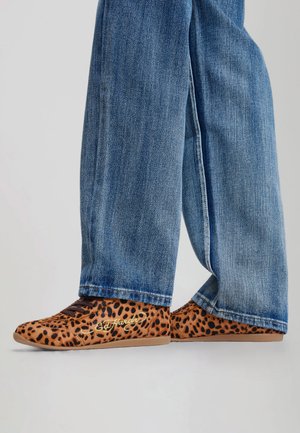 Persoon die wijde blauwe spijkerbroek draagt en platte schoenen met luipaardprint met geborduurd logo aan de zijkant, staand op een lichte achtergrond.