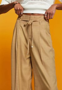Beige wide-leg trousers with a textured fabric, featuring a tie waist and front pleats. Set against an orange background.