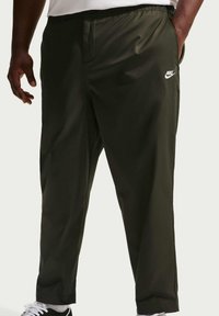 Homme portant un pantalon de survêtement Nike noir avec logo blanc, debout avec une main dans la poche et des baskets blanches visibles.