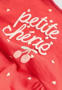 Sarkans kokvilnas audums ar baltu tekstu "petite chérie", rozā un zaļām ķiršu grafikām un mazām krēmkrāsas sirdīm, kas izkaisītas apkārt.