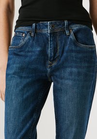 Sieviete, kas valkā tumši zilas džinsa bikses un melnu topu, rādot vidukļa un augšstilbu apvidu ar redzamām šuvēm un kabatām.