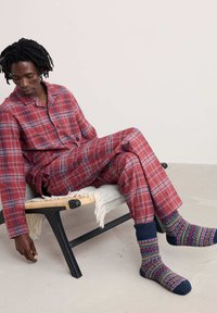 Henkilö punaisissa ruudullisissa pyjamahousuissa ja kuvioiduissa sukissa istuu matalalla tuolilla, katsoo alas ja koskettaa lattiaa yhdellä kädellä.