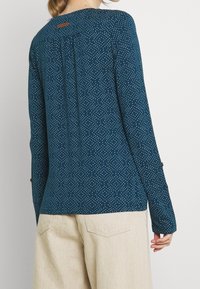 Tyrkysová blůza s bílými puntíky. Dlouhé rukávy s manžetami na knoflíky. Šev na zadní straně a malá hnědá etiketa na límci.