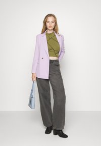 Blazer lilás, top cropped verde-azeitona com padrão geométrico preto, jeans cinza de perna larga de cintura alta, botas de salto pretas e uma bolsa azul-claro.
