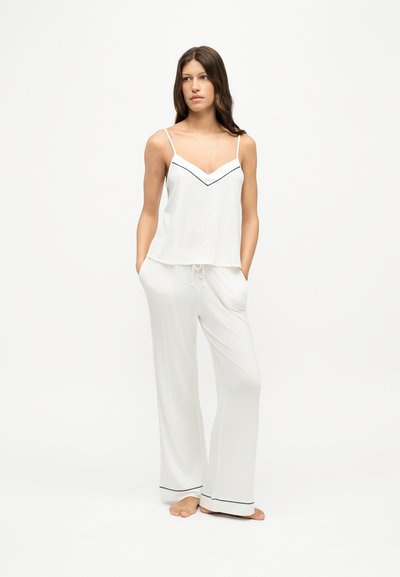 Femme debout pieds nus portant un ensemble de pyjama blanc avec fines bretelles et passepoil noir sur le haut et les ourlets du pantalon, mains dans les poches, sur fond uni.