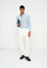 Világoskék, gombos ing zsebbel, fehér nadrág, fekete vastag talpú cipő, fekete táska a kezében. Egyszínű fehér háttér előtt.