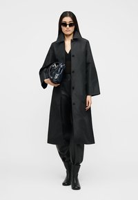 Γυναίκα που φοράει μακρύ μαύρο παλτό, μαύρο παντελόνι, μαύρες μπότες, σκούρα γυαλιά ηλίου, κρατώντας μια τσάντα χρώματος ναυτικού μπλε σε απλό ανοιχτό φόντο.