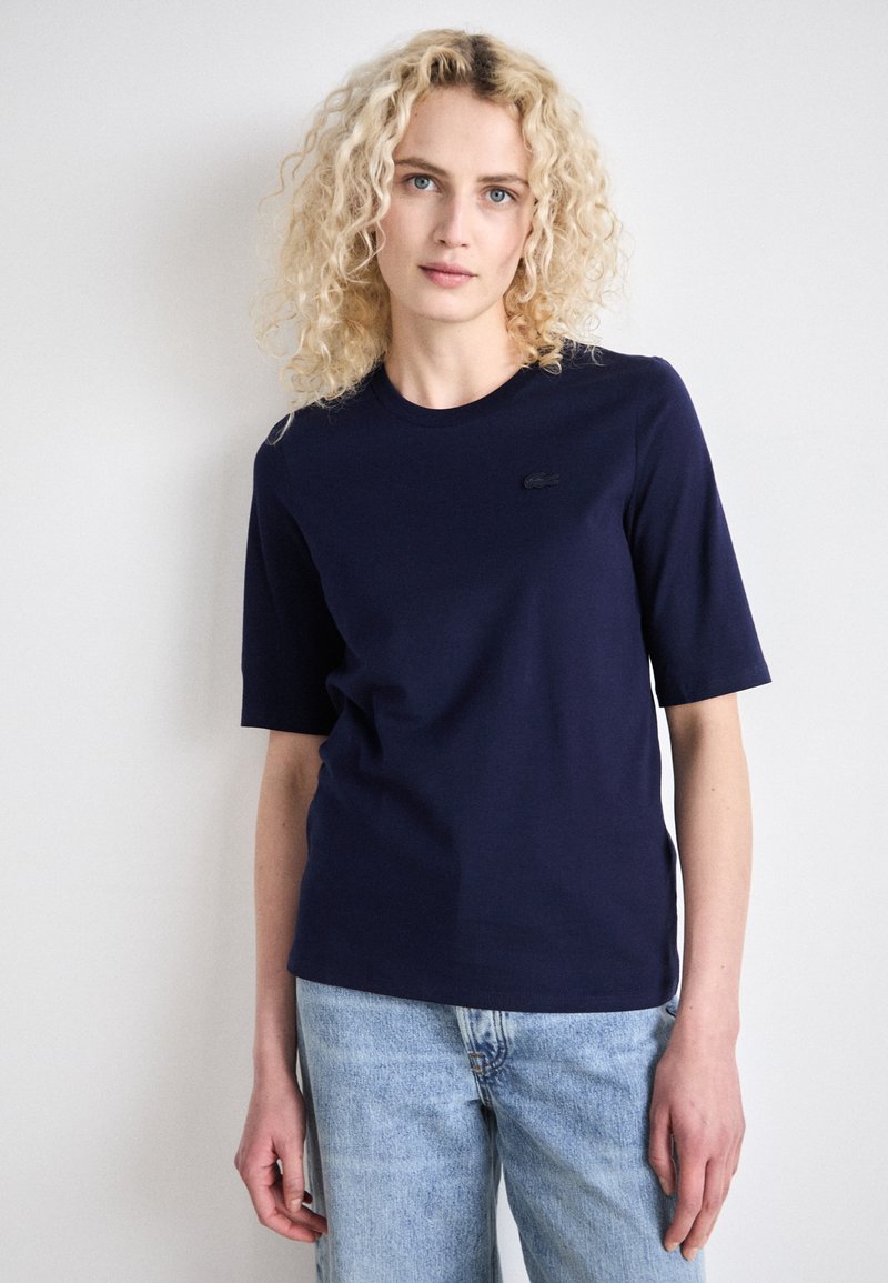 T-shirt de algodão azul-marinho de manga curta com um decote redondo e um pequeno logótipo bordado no peito, combinado com jeans de perna larga em azul claro.