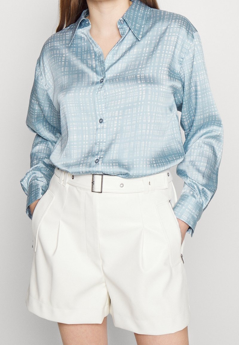 Chemise en soie bleu clair à motifs avec un devant à boutons et un col, associée à un short blanc taille haute avec des poches latérales.