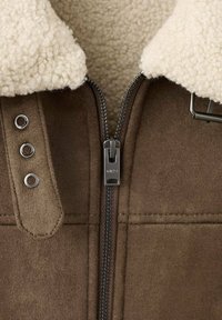 Giacca in suede marrone con colletto in shearling crema, zip centrale e hardware in metallo. Presenta tre bottoni a pressione decorativi sulla spalla.