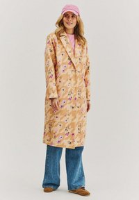 Brožový trenčkot s květinovými a geometrickými vzory, se dvěma knoflíky a límcem, nošený přes růžový svetr a modré zvonové džíny, doplněný růžovou čepicí.